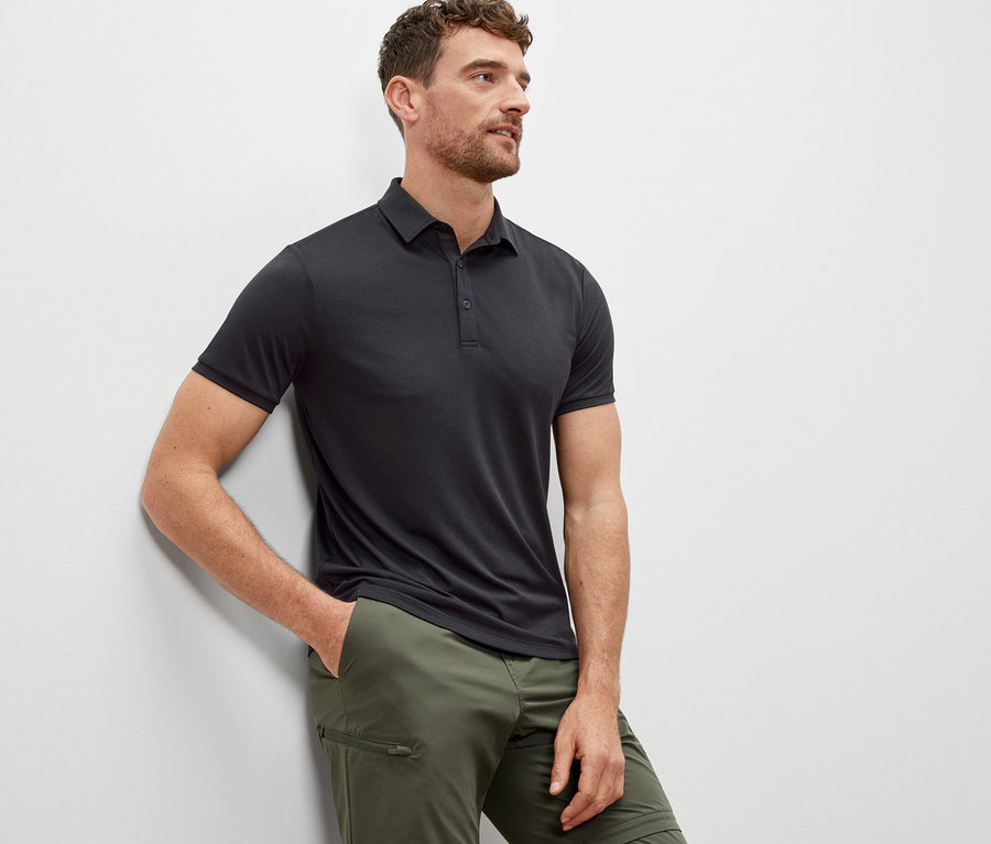 Mann lehnt an einer Wand und trägt ein anthrazitfarbenes Funktions-Poloshirt.