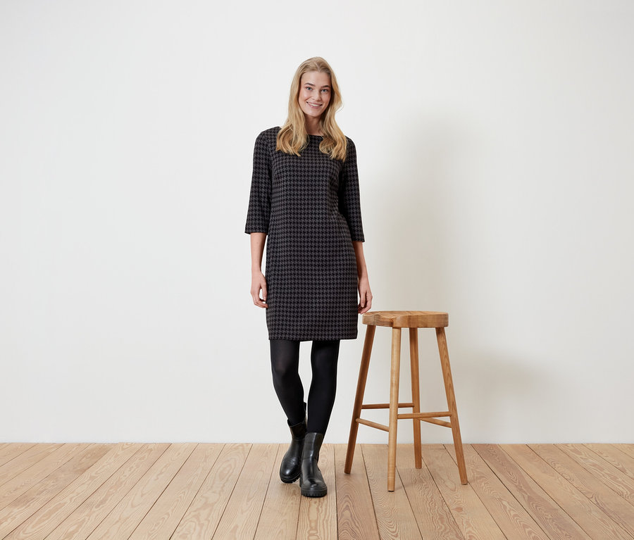 Frau steht neben einem Holzhocker und trägt ein Hahnentrittkleid, schwarze Leggings und Stiefel.