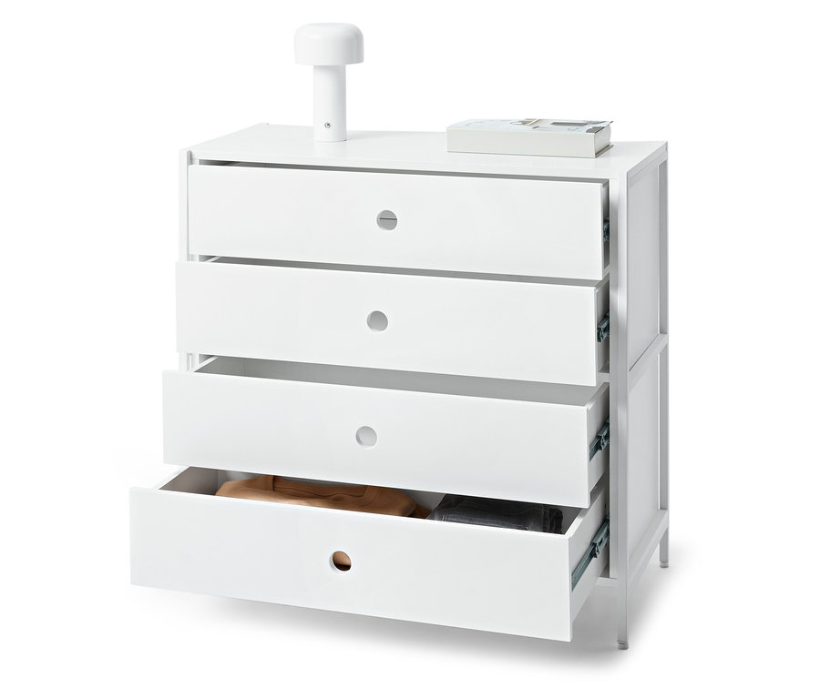 Weiße Kommode mit vier Schubladen, von denen eine geöffnet ist, darauf liegen eine Lampe und ein Buch.