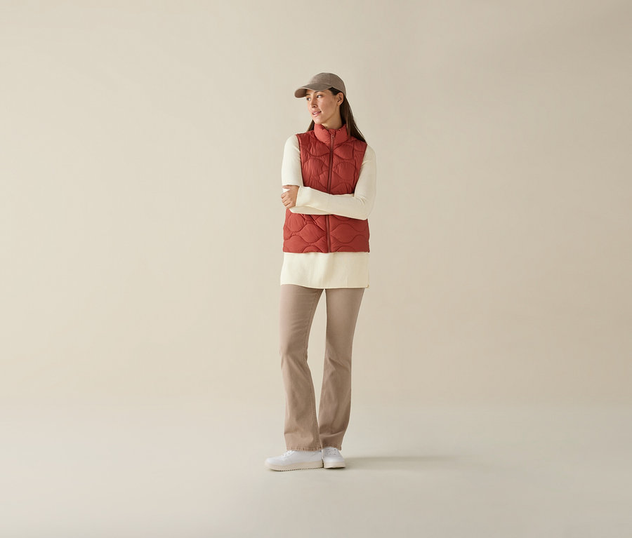 Frau mit verschränkten Armen trägt eine Steppweste, ein cremefarbenes Oberteil, beige Hosen und eine Kappe.