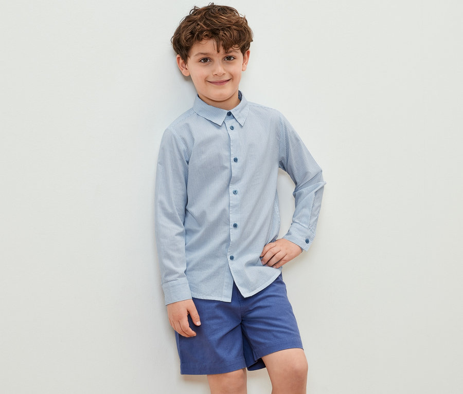 Junge posiert in einem Kinder-Hemd und blauen Shorts.