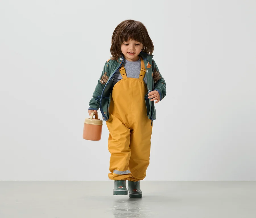 Kleinkind in gelber Latzhose, grüner Jacke und Gummistiefeln hält eine orangefarbene Lunchbox in der Hand.
