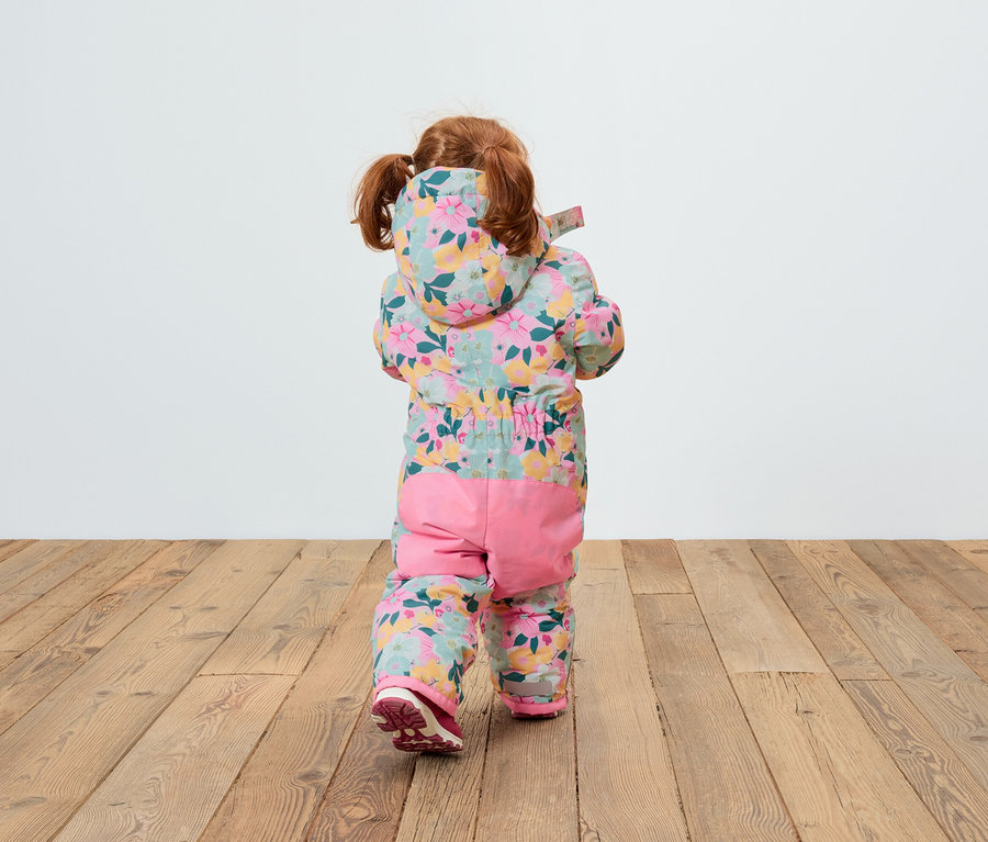 Rückansicht eines Kindes mit roten Haaren in einem geblümten Overall mit Kapuze und rosa Hosen, das auf einem Holzboden steht.