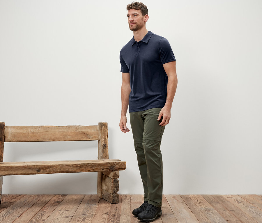 Mann steht neben einer Holzbank und trägt ein blaues Poloshirt mit Merinowolle.