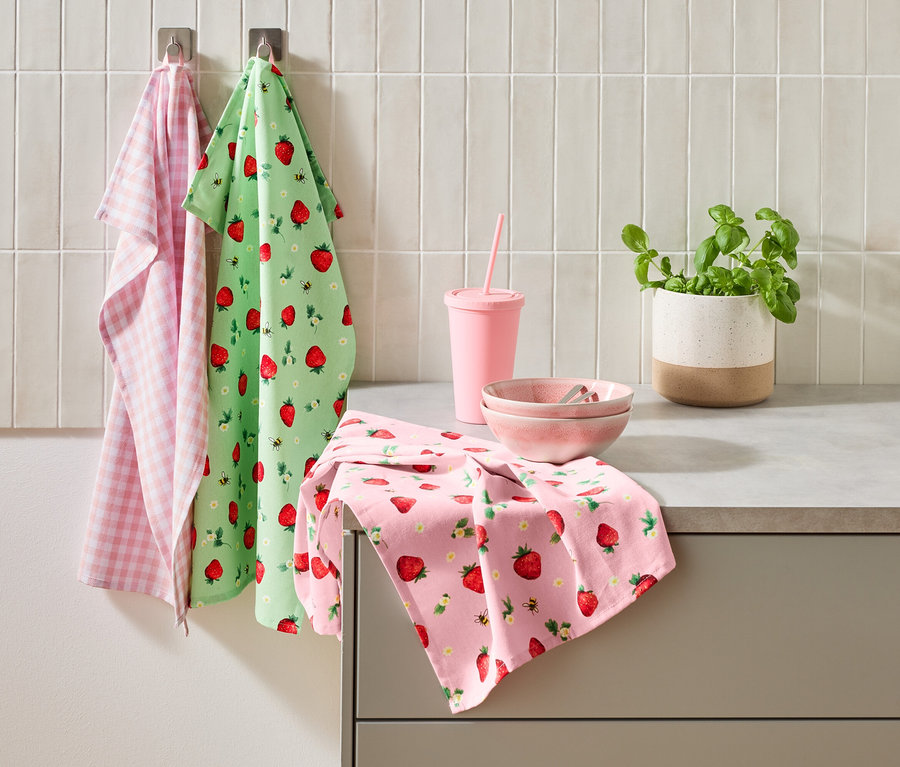 Drei Küchentücher, ein rosa Becher mit Strohhalm, zwei rosa Schalen und ein Topf Basilikum stehen auf einer Küchentheke.