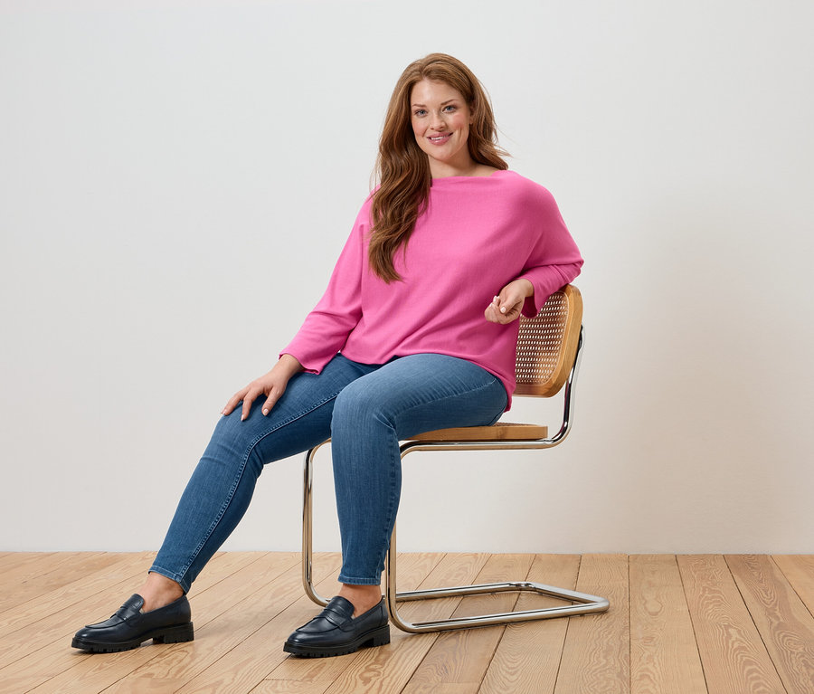 Eine Frau sitzt auf einem Stuhl und trägt einen pinkfarbenen Pullover, blaue Jeans und schwarze Loafer.