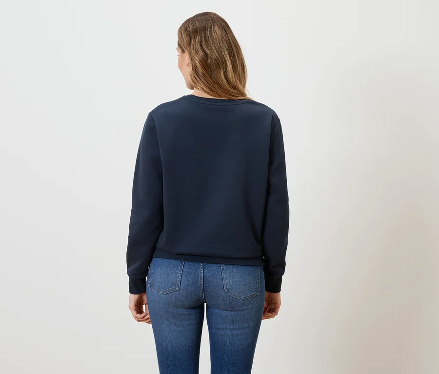 Rückansicht einer Frau mit langen, gewellten, hellbraunen Haaren, die einen marineblauen Pullover und Jeans trägt.