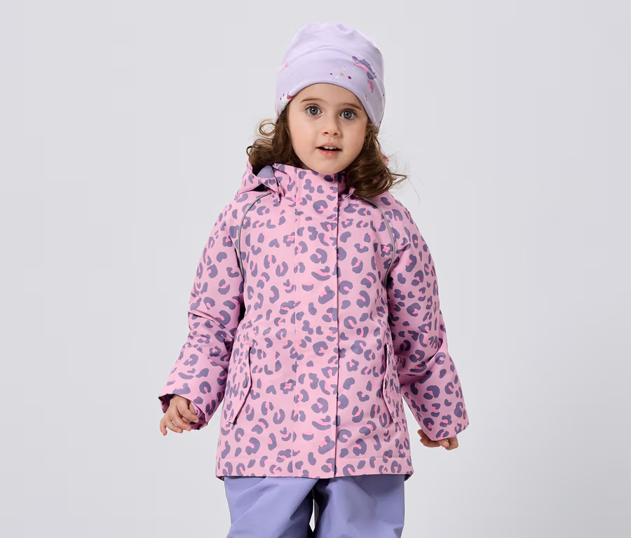 Ein kleines Mädchen mit lockigem Haar trägt eine rosa-lila Jacke mit Leopardenmuster, eine passende Mütze und lila Hosen vor einem weißen Hintergrund.