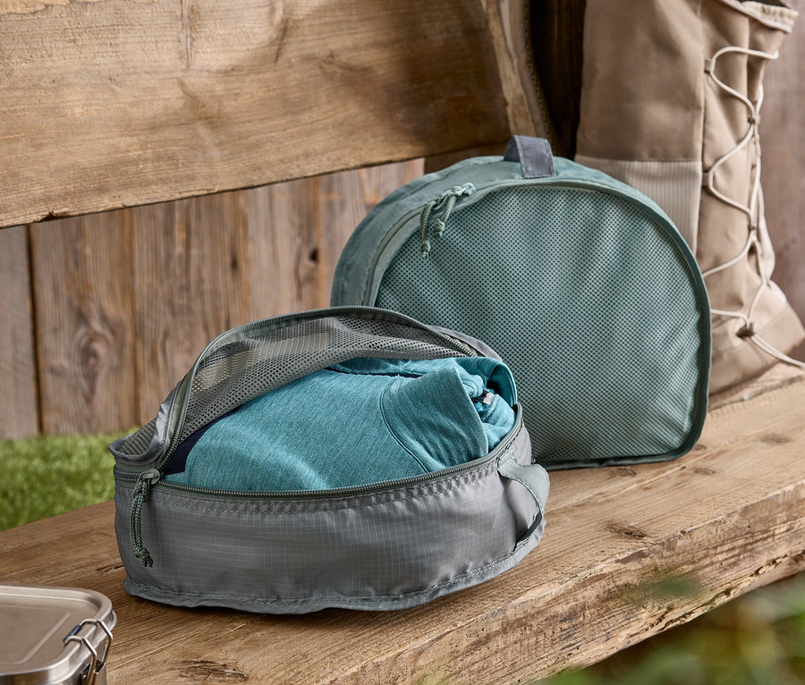 Grauer Packwürfel mit blauem T-Shirt teilweise geöffnet auf einer Holzbank. Im Hintergrund ein grüner Packwürfel und ein Rucksack.