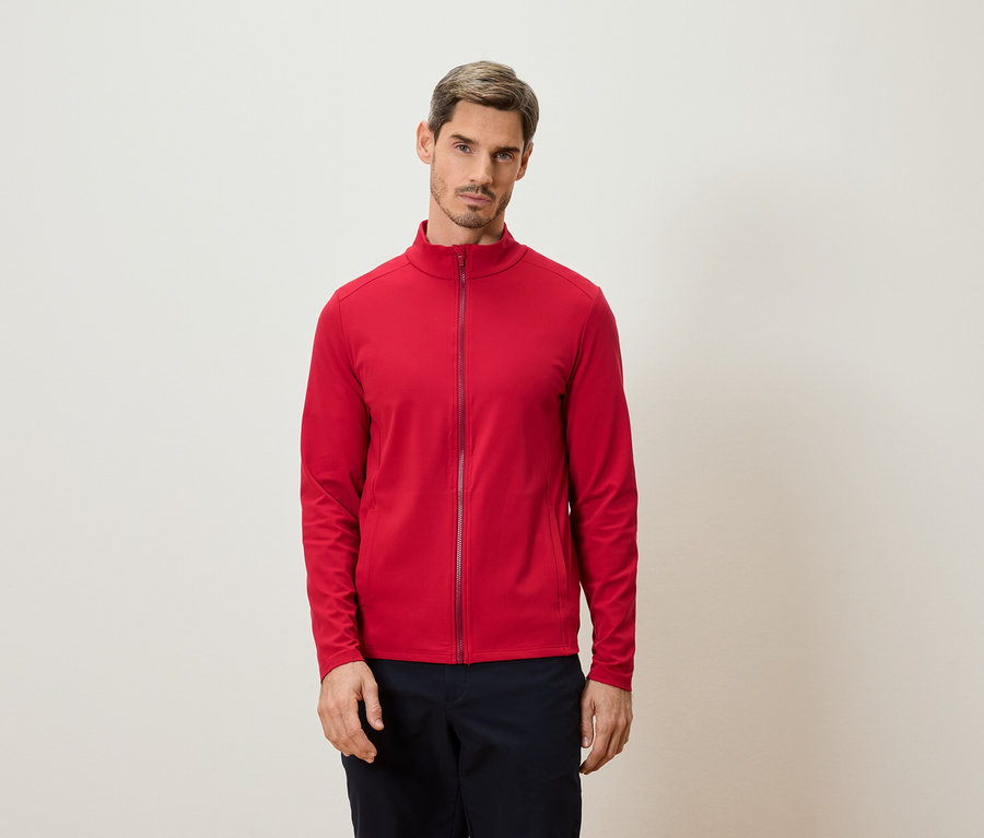Mann steht vor einer Wand und trägt eine rote Jacke mit Reißverschluss.
