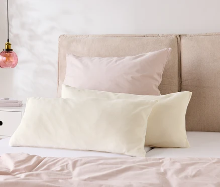 Ein Bett mit hellrosa Bettwäsche und zwei cremefarbenen Kissen liegt vor einem beigen Kopfteil. Auf dem Nachttisch steht eine Lampe.