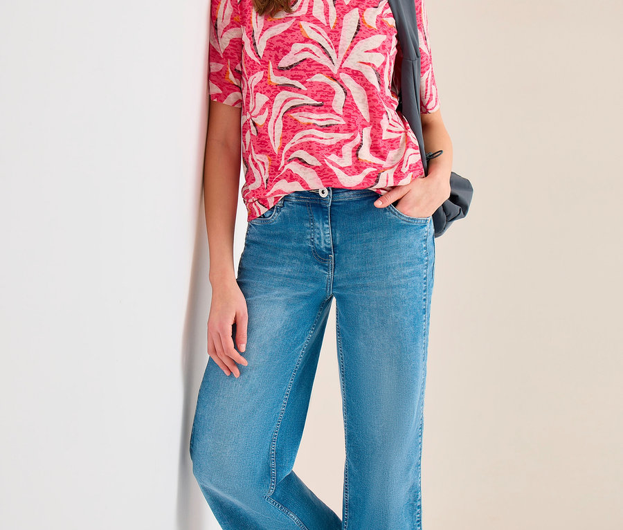 Frau lehnt an einer weißen Wand, trägt ein rosa CECIL Kurzarmshirt mit weißem Muster, weite blaue Jeans und eine dunkle Umhängetasche.