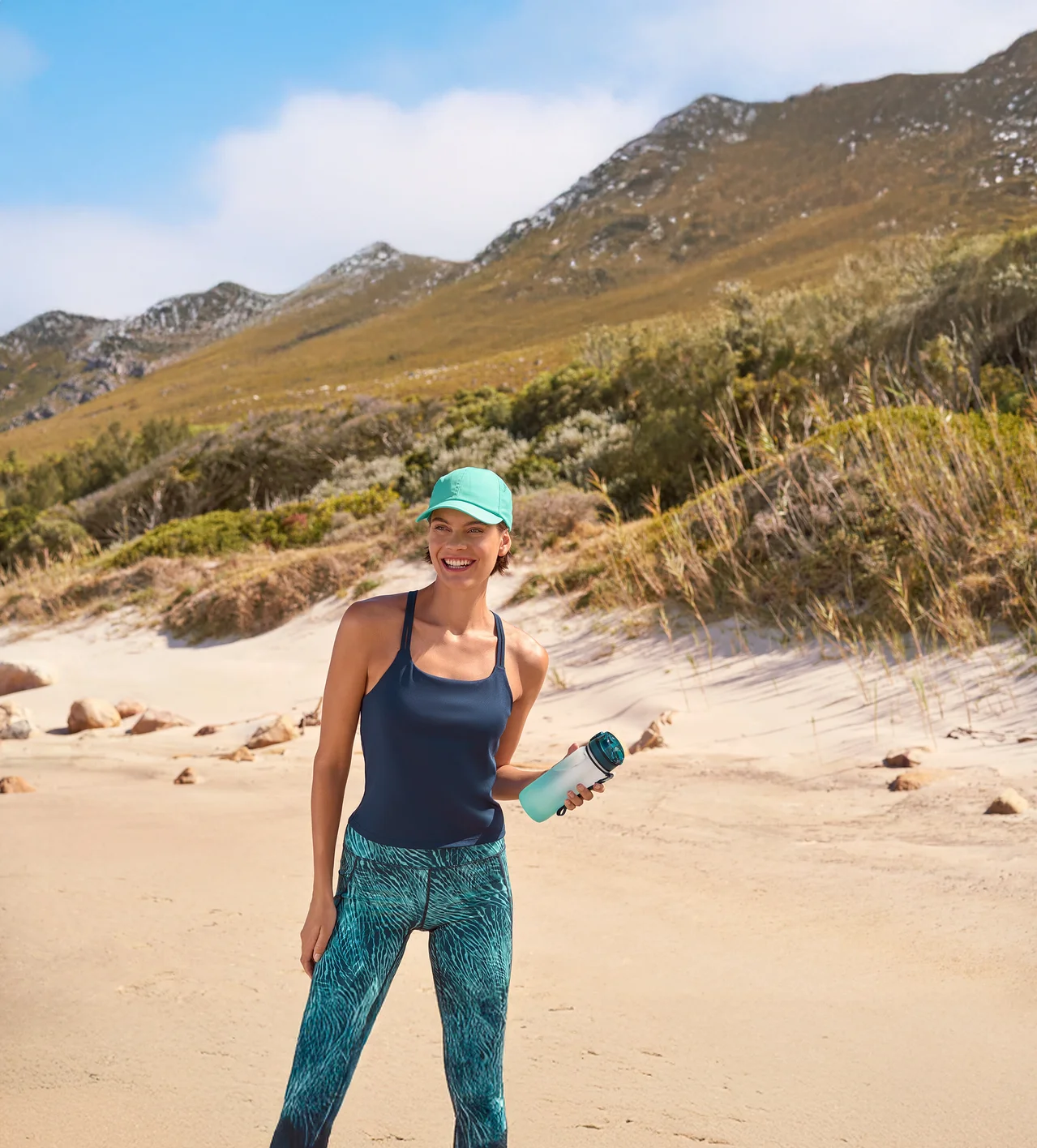 Lächelnde Frau in türkisener Sport-Kappe, dunkelblauem Yogatop mit integriertem BH und Sportradler »Active Power« mit Alloverprint, die eine Trinkflasche mit Kühlakku am Strand hält. Im Hintergrund sind Berge zu sehen.