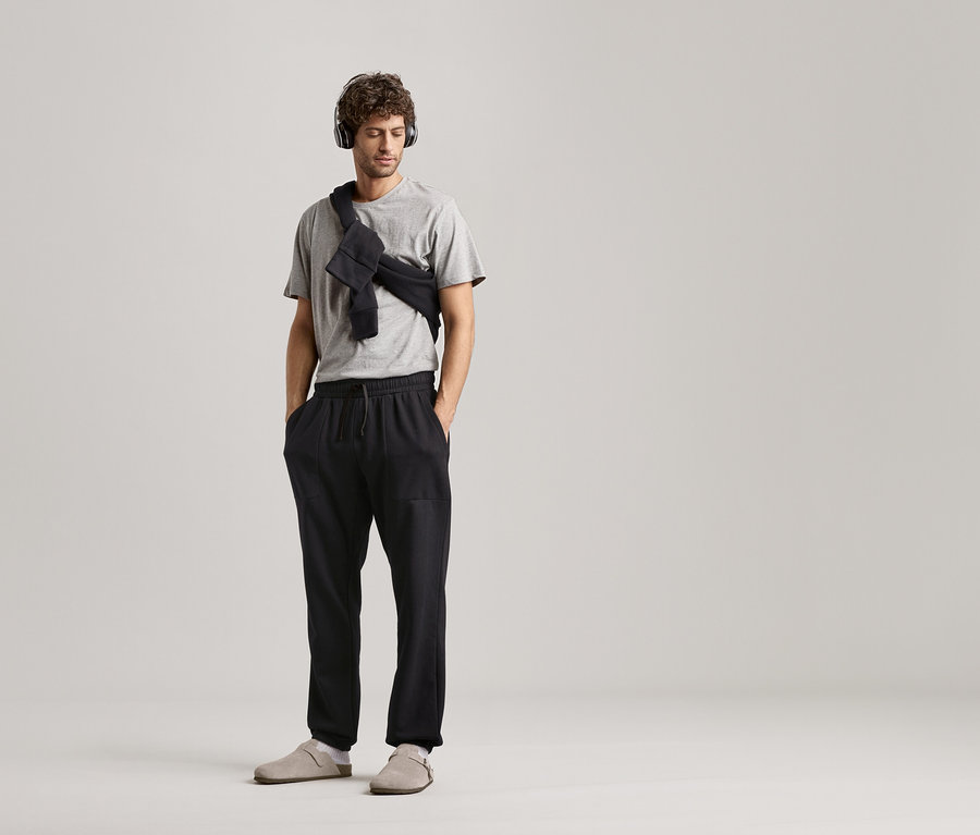 Mann mit Kopfhörern, grauem T-Shirt und schwarzer Sweathose vor neutralem Hintergrund.