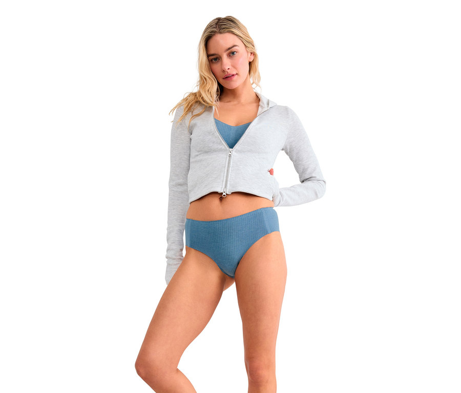 Eine Frau mit blonden Haaren steht vor weißem Hintergrund und trägt einen hellgrauen, bauchfreien Kapuzenpullover mit Reißverschluss, einen blauen Bralette und blaue gerippte Slips. Ihre Hände sind hinter ihrem Rücken.