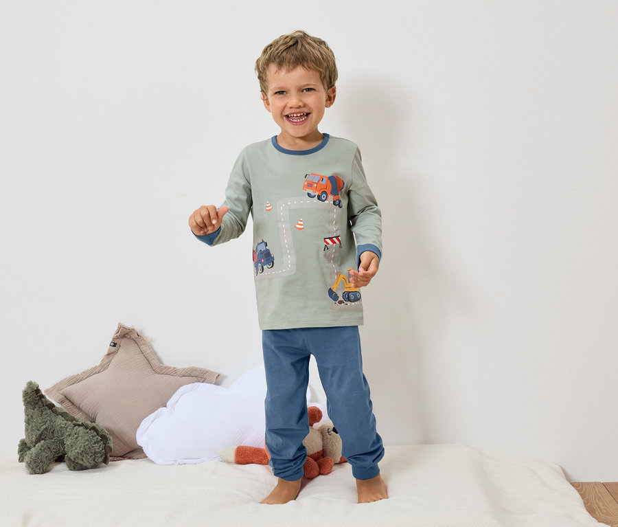 Ein lächelnder Junge steht barfuß auf einer weißen Oberfläche und trägt einen grauen Pyjama mit blauen Bündchen und einem Bagger- und Betonmischer-Motiv.