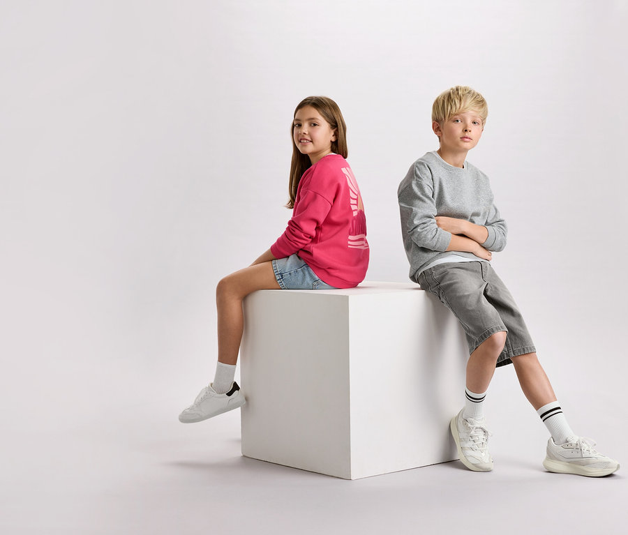 Mädchen mit braunen Haaren in einem pinken Pullover und Jeansshorts sitzt neben einem Jungen mit blonden Haaren in einem grauen Pullover und Shorts auf einem weißen Würfel.