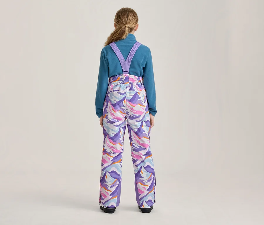 Eine Person von hinten, die ein blaues Langarmshirt und eine bunte Skihose mit lila Trägern trägt. Die Hose hat ein abstraktes Muster in Lila, Pink, Orange und Weiß.