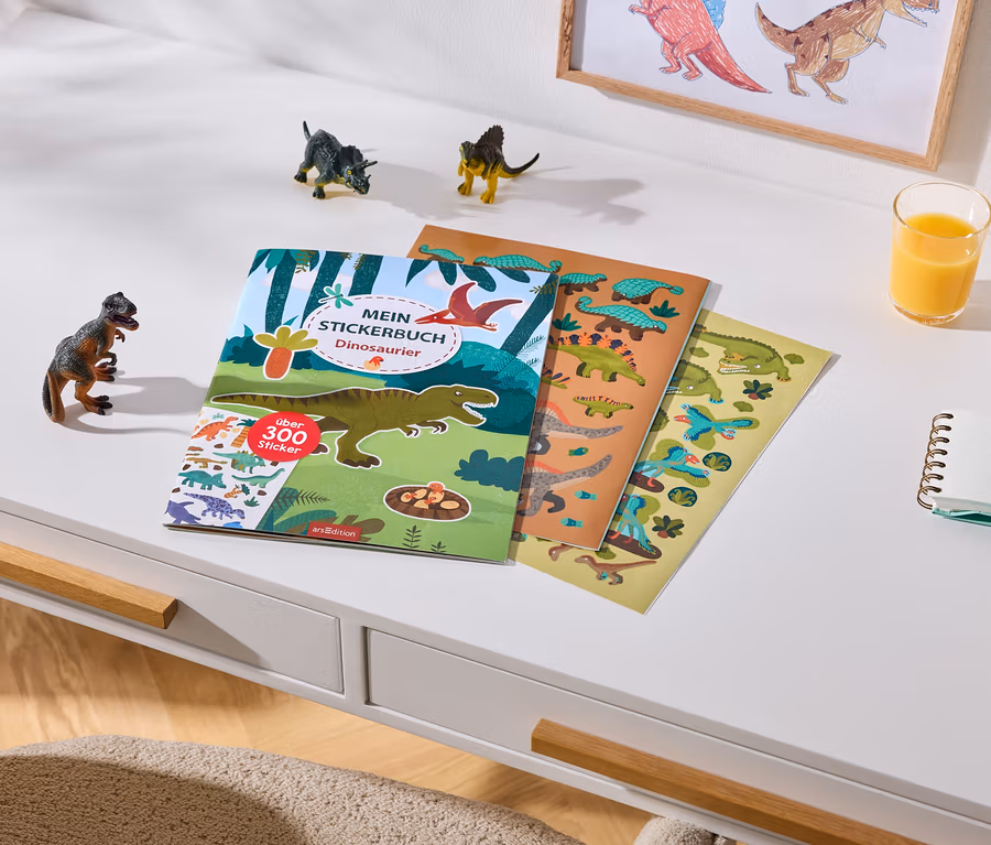 Ein weißer Schreibtisch mit einem Dinosaurier-Stickerbuch, das über 300 Sticker enthält, und zwei Stickerbögen. Daneben stehen drei kleine Dinosaurierfiguren, ein Glas Orangensaft, ein Notizbuch und zwei Buntstifte. Im Hintergrund hängt ein gerahmtes Dinosaurierbild an der Wand.