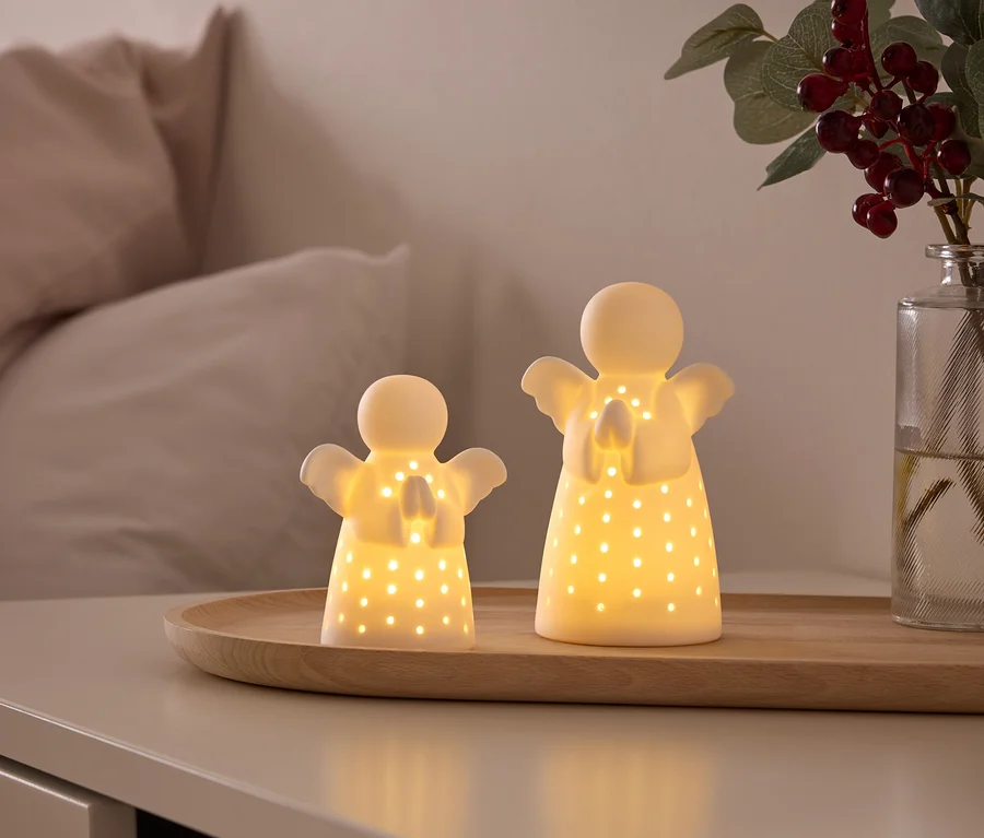 Zwei leuchtende, weiße Engelsfiguren mit ausgestanzten Löchern stehen auf einem Holztablett auf einem Nachttisch. Im Hintergrund sind ein Bett und eine Vase mit roten Beeren zu sehen.