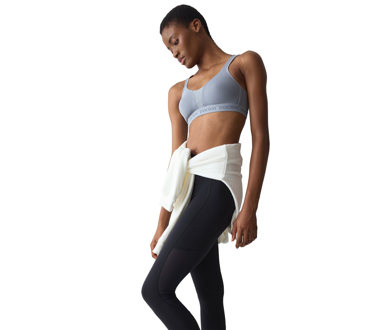Frau mit kurzen dunklen Haaren trägt einen grauen Sport-BH und schwarze Leggings, ein weißes Oberteil ist um ihre Taille gebunden, sie blickt nach unten.
