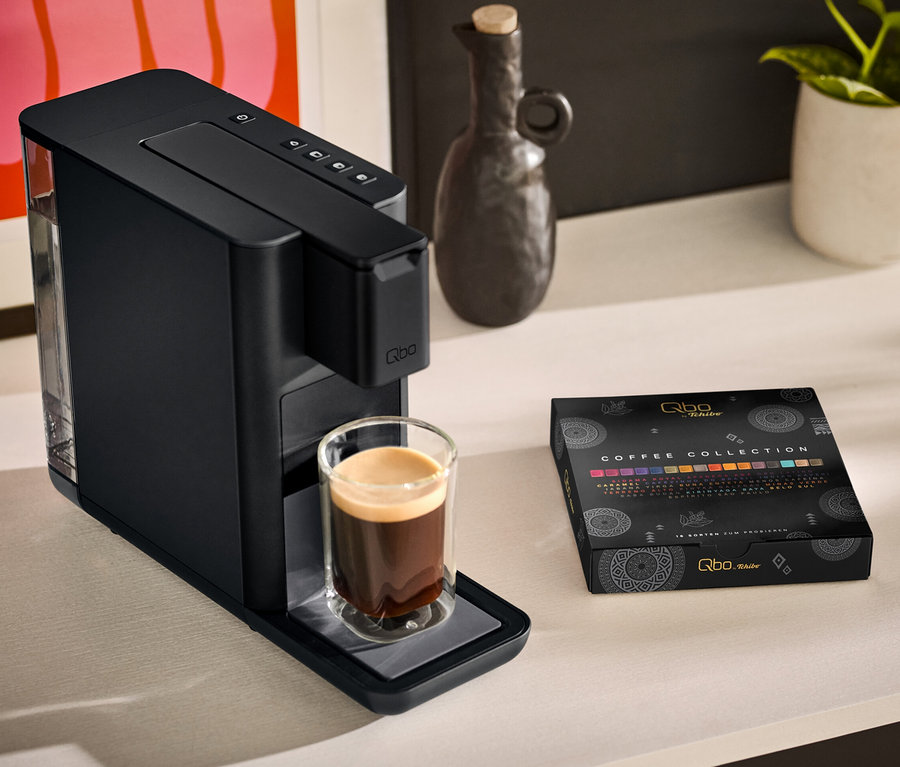 Eine Qbo ICONIC Dark Chrome Kaffeemaschine mit einer Tasse Kaffee daneben und eine Qbo Collection - 16 Kapseln Packung stehen auf einem Tisch.