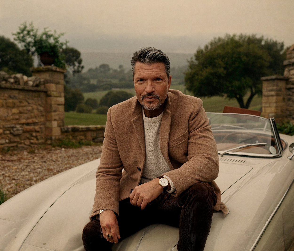 Ein Mann mit grauem Haar und Bart sitzt auf der Motorhaube eines hellen Cabrios. Er trägt einen beigefarbenen Mantel, einen cremefarbenen Pullover und eine goldfarbene Kienzle Uhr »Meister Klassik« am Handgelenk. Im Hintergrund sind eine Steinmauer und grüne Felder zu sehen.