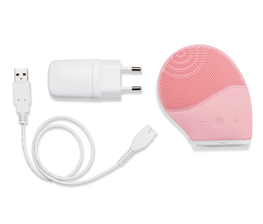 Eine elektrische Silikon-Gesichtsreinigungsbürste in Rosa liegt mit Ladekabel und Adapter auf weißem Grund.