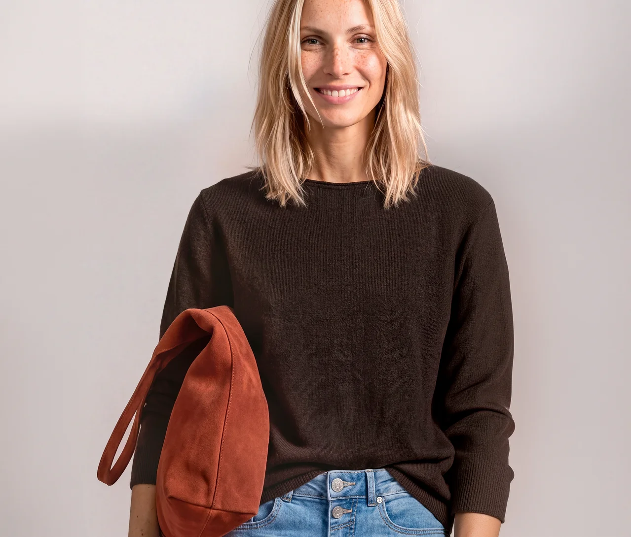 Lächelnde Frau trägt braunen Street One Strickpullover und hellblaue Jeans, hält eine rotbraune Tasche.