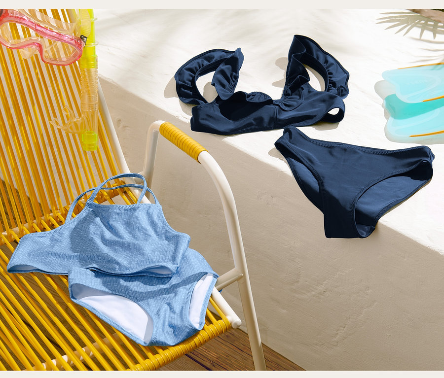 Ein blaues Bikini-Set liegt mit Schnorchel und Flossen auf einem gelben Strandstuhl und einer weißen Wand.