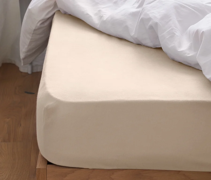 Ein beiges Biber-Spannbettlaken auf einem Holzbett mit weißen Laken und Kissen.