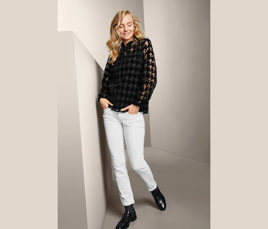 Blonde Frau posiert in Mesh-Bluse mit Hahnentrittmuster, weißen Jeans und schwarzen Schuhen.