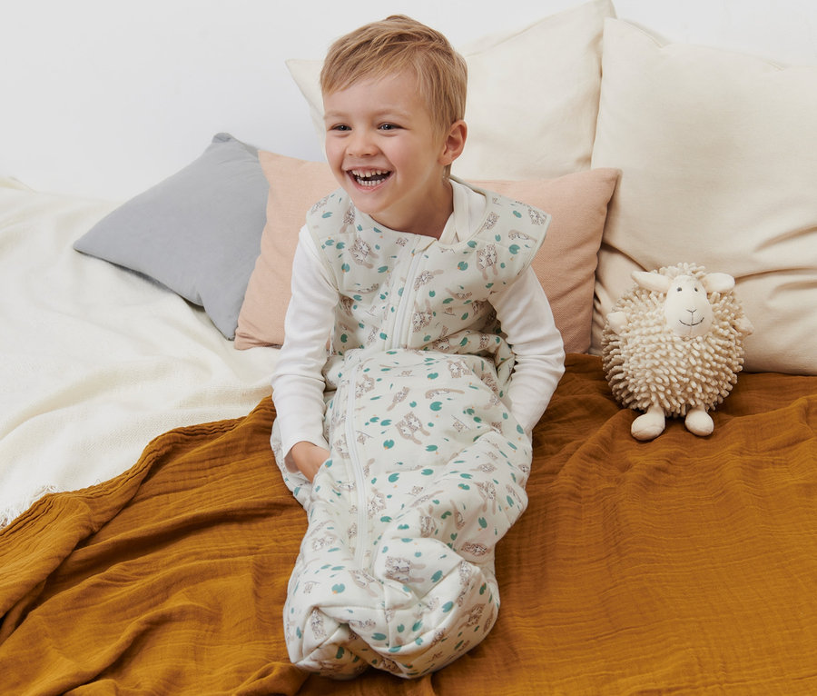 Lächelnder Junge sitzt in Schlafsack 2,5 TOG, 110 cm, mit Otter-Print auf dem Bett.