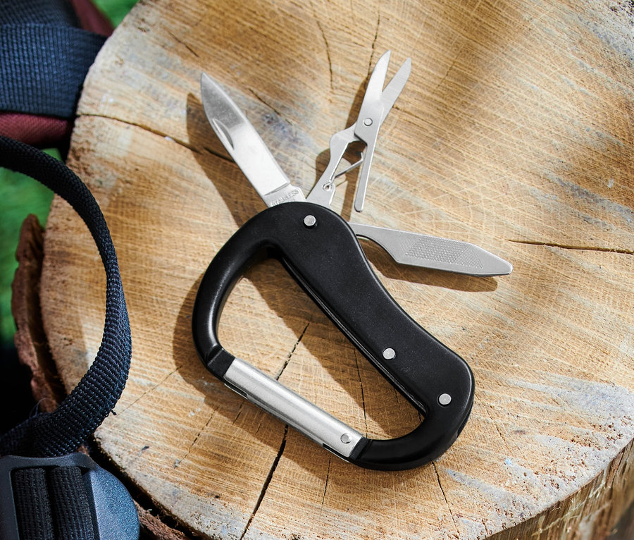 Ein 5-in-1-Karabiner-Multitool liegt auf einem Holzscheit neben einem Rucksack.