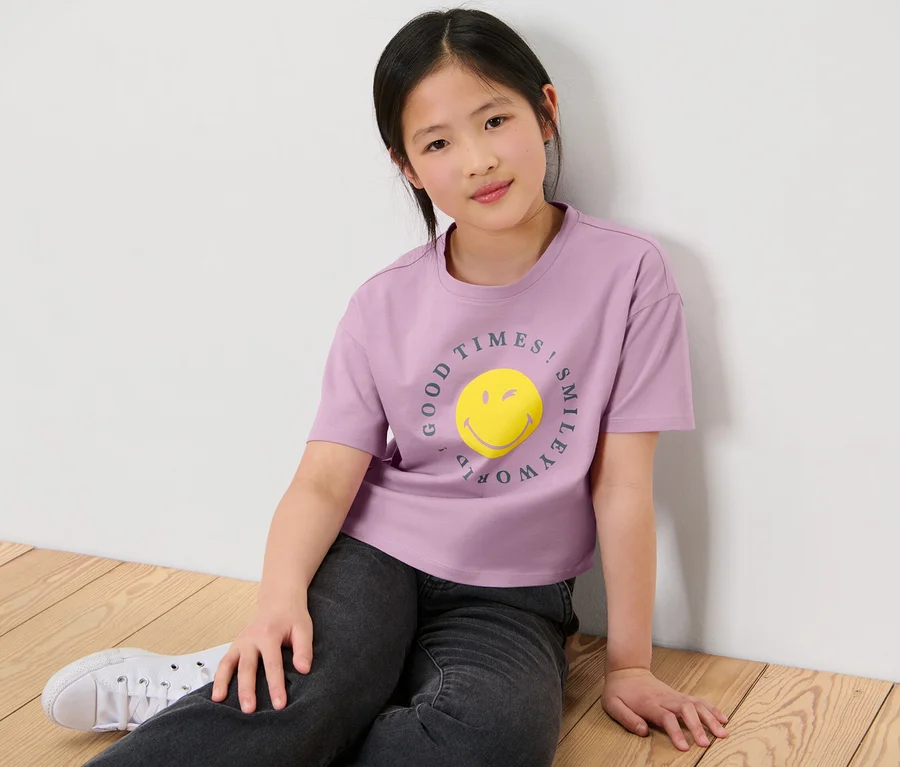 Mädchen sitzt auf dem Boden und trägt ein lila T-Shirt mit »Smiley« Aufdruck, 2 Kinder-T-Shirts.
