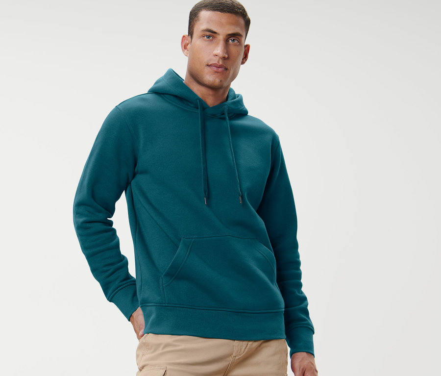 Mann posiert vor hellem Hintergrund in petrolfarbenem Hoodie.