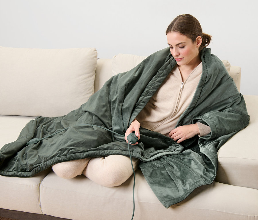 Frau sitzt auf Sofa und ist mit einer Heizdecke zugedeckt, sie hält eine Fernbedienung in der Hand.