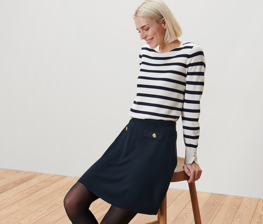 Blonde Frau sitzt auf einem Hocker und trägt einen Strickrock mit Zier-Knöpfen und ein gestreiftes Oberteil.