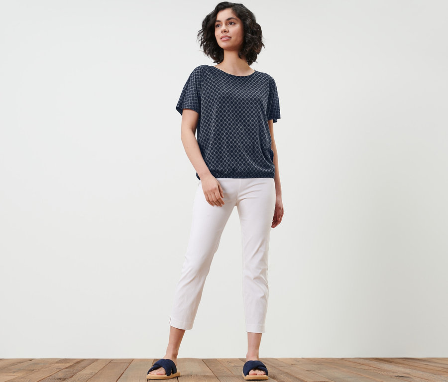 Frau posiert in einem bedruckten Blusenshirt, weißen Hosen und Pantoletten.