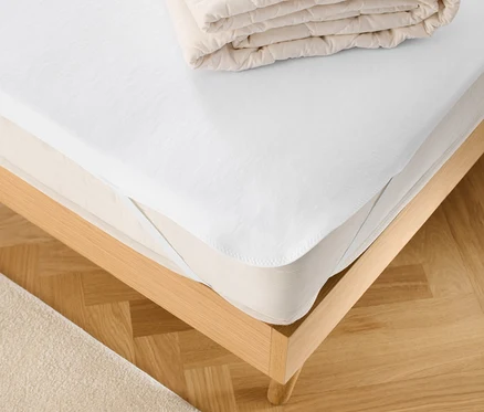 Auf einem Bett liegen ein weißer Matratzenschoner und eine gefaltete Decke.
