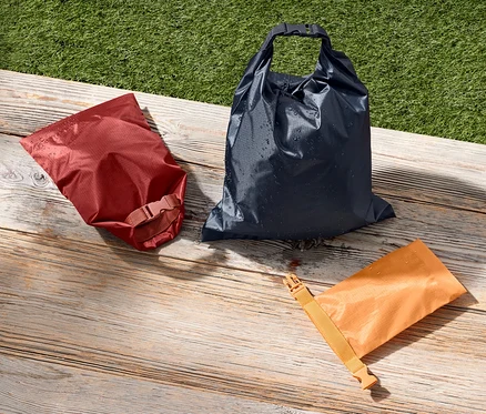 Drei wasserfeste Packsäcke in Rot, Blau und Orange liegen auf einer Holzoberfläche neben einem grünen Rasen.