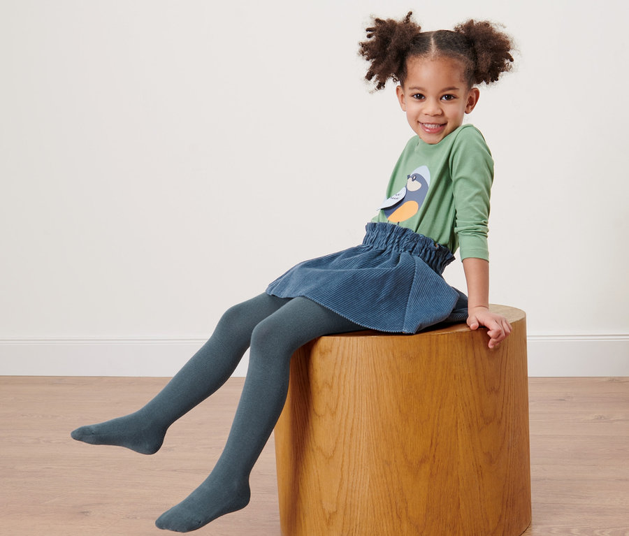 Ein lächelndes Mädchen sitzt auf einem Holzhocker und trägt eine blaue Kinderstrumpfhose.