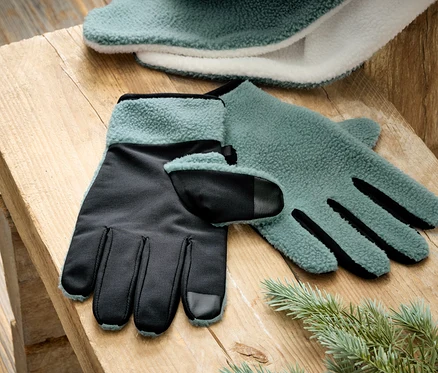 Ein Paar türkis-schwarze Fleecehandschuhe liegen auf einem Holztisch neben einem Tannenzweig.