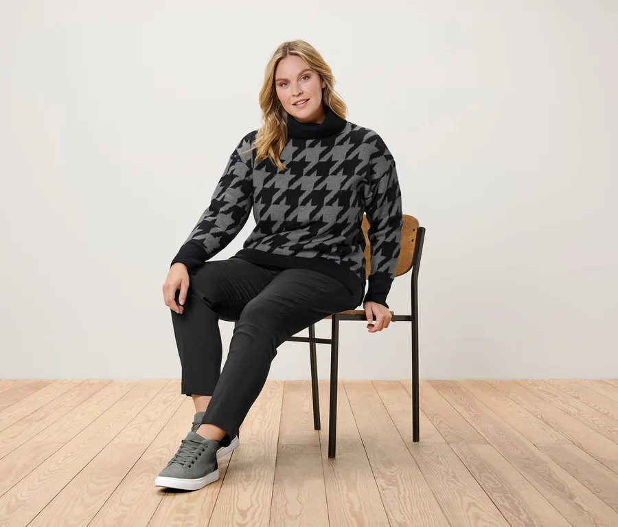 Frau sitzt auf einem Stuhl und trägt einen Hahnentrittpullover, eine schwarze Hose und graue Turnschuhe.