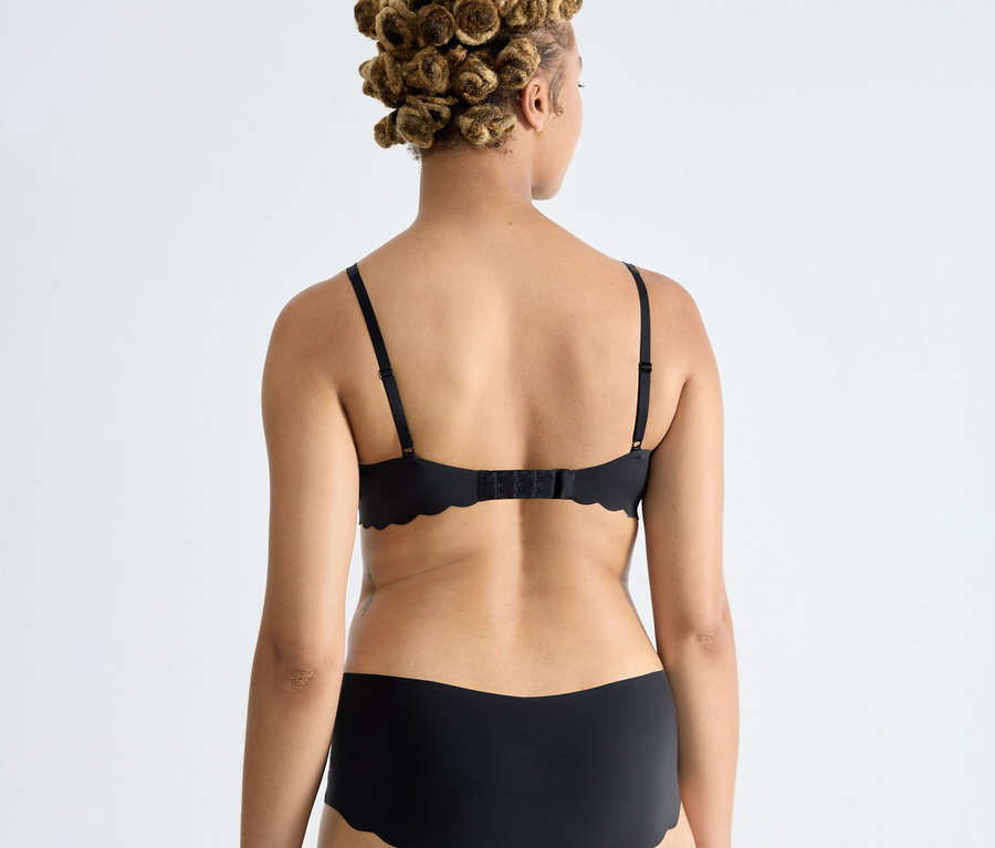 Frau von hinten, trägt einen schwarzen sloggi ZERO Microfibre 2.0 Soft bra und passende schwarze Unterwäsche mit Wellenkanten.