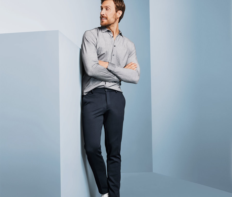 Mann in grauem Hemd und dunkelblauen Jersey-Chinos, der an einer Wand lehnt.