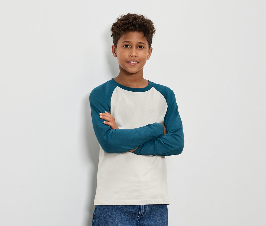 Junge mit braunem lockigem Haar steht mit verschränkten Armen und trägt ein blaugrünes Langarmshirt und Jeans.