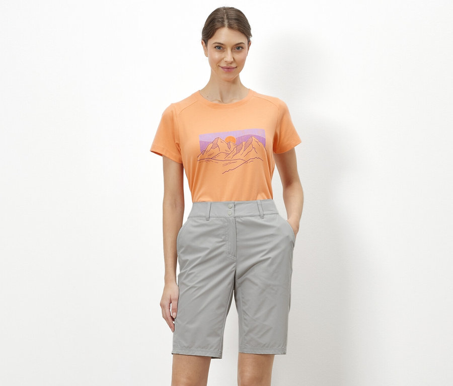Eine Frau steht vor einem weißen Hintergrund und trägt ein orangefarbenes T-Shirt mit einer Abbildung von Bergen und graue Rad-und-Wandershorts »Pro«.