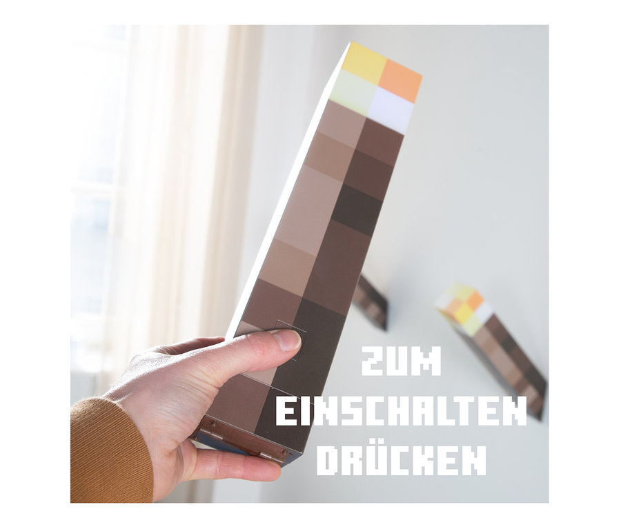 Eine Hand hält eine MINECRAFT LED Dekoleuchte, Fackel. Im Hintergrund ist eine weitere Fackel an der Wand befestigt.