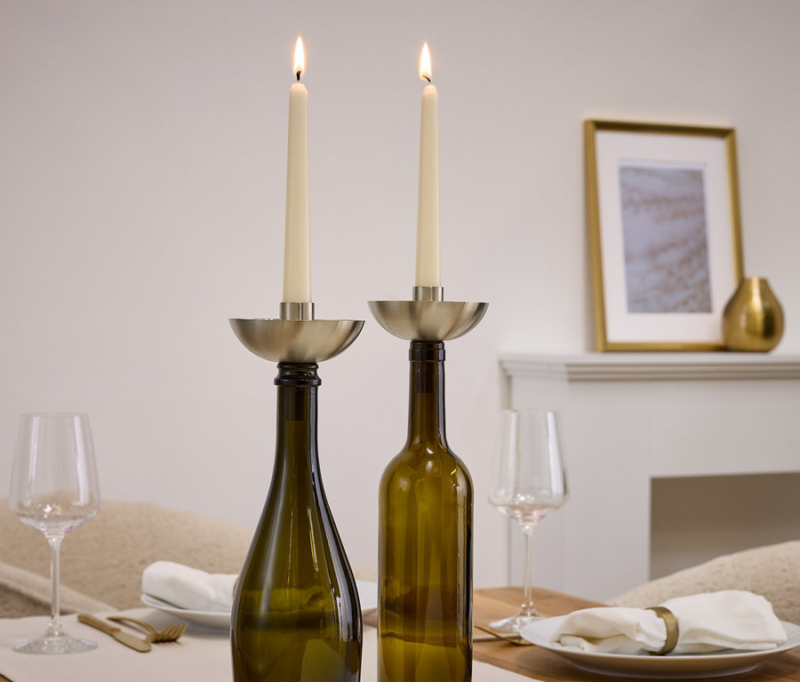 Zwei brennende Kerzen in silbernen Kerzenhaltern auf grünen Flaschen, daneben Gläser und Teller.